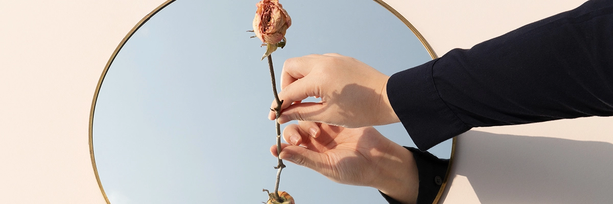 Mão de mulher segurando uma rosa em frente um espelho que reflete a imagem da flor.