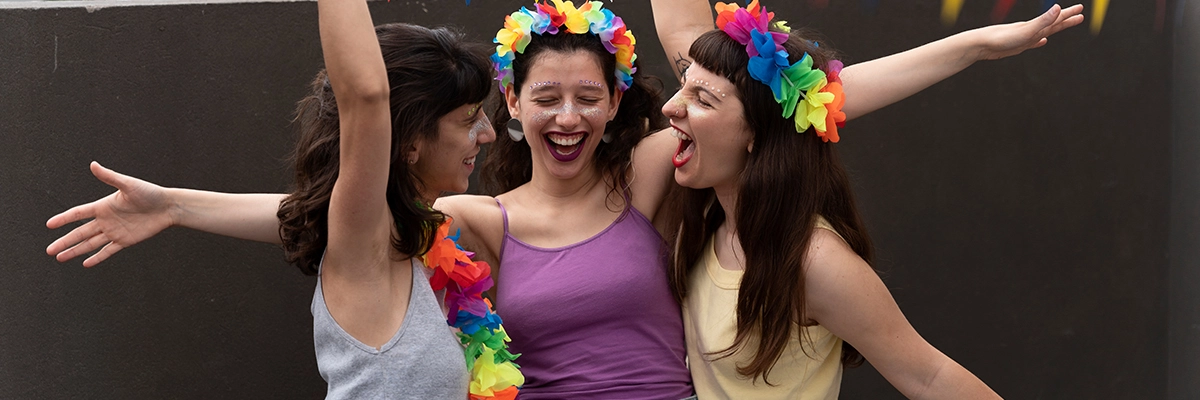 Três mulheres felizes com trajes de carnaval dançando e festejando juntas sorrindo.